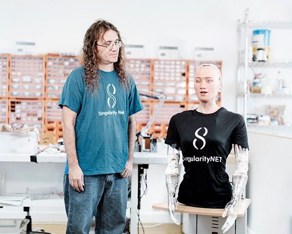 Dr Ben Goertzel stands beside Sophia's head and torso, which sits on a plinth in a workroom with shelves and drawers of parts and tools on the walls and tables. Both wear T-shirts bearing SingularityNET's logo.