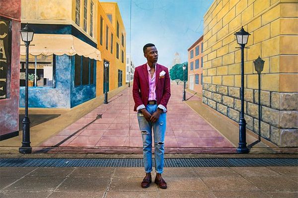 South African fashion blogger Ofentse Lewis wears a red blazer and jeans and stands in front of a painted mural showing a smart street scene.
