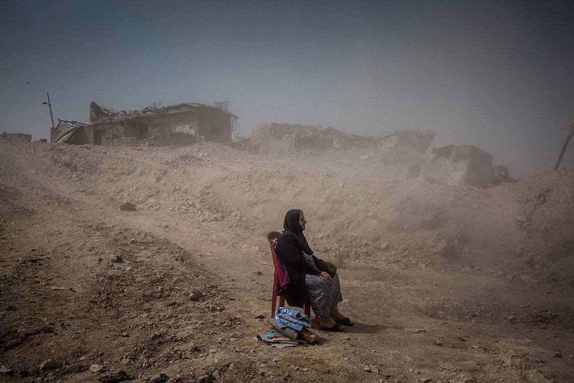 An elderly Iraqi lady sits on a dining chair in the middle of a dusty rural road with collapsed houses at the side.