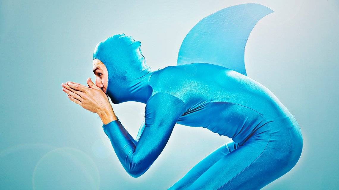 A man wears a shiny, metallic blue all-in-one outfit with a head covering and a shark fin on his back. He is poised as if to dive.
