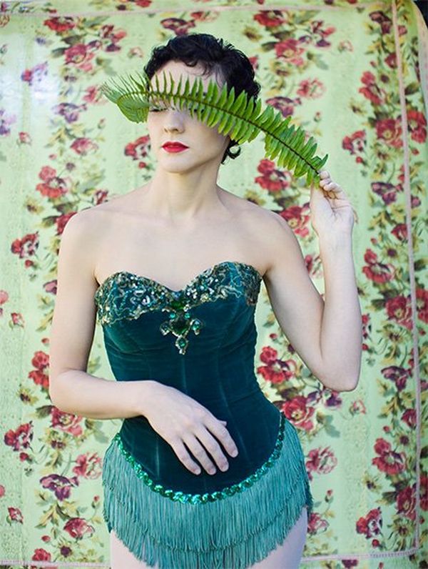 A woman in an emerald-coloured velvet corset trimmed with tassels and beads holds a fern frond across her eyes.