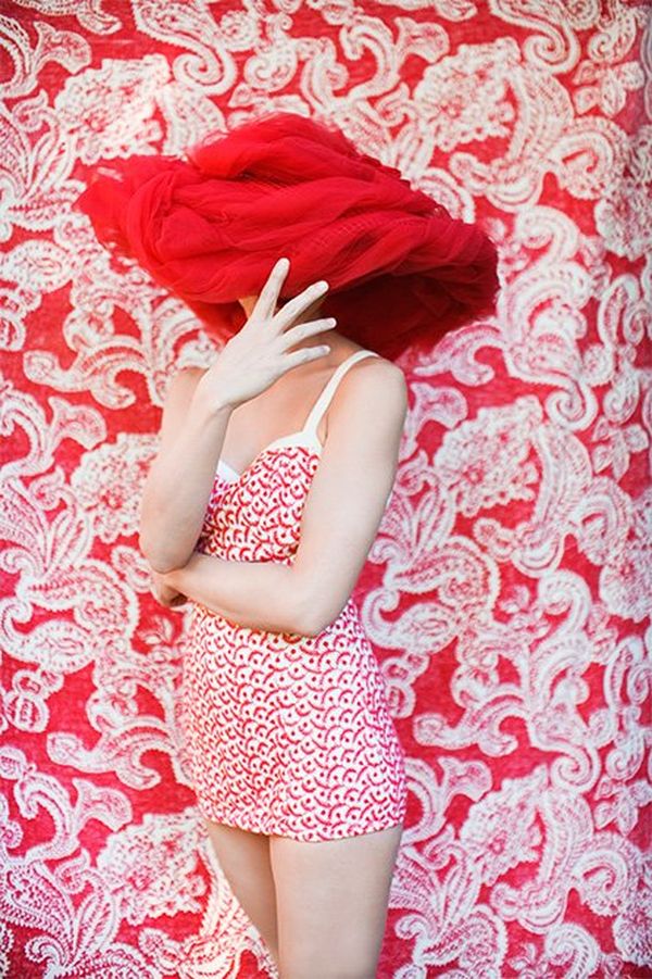A woman in a red-and-white retro-style swimsuit wears an oversized hat on the head, covering her eyes, and stands in front of a red-and-white patterned wall.