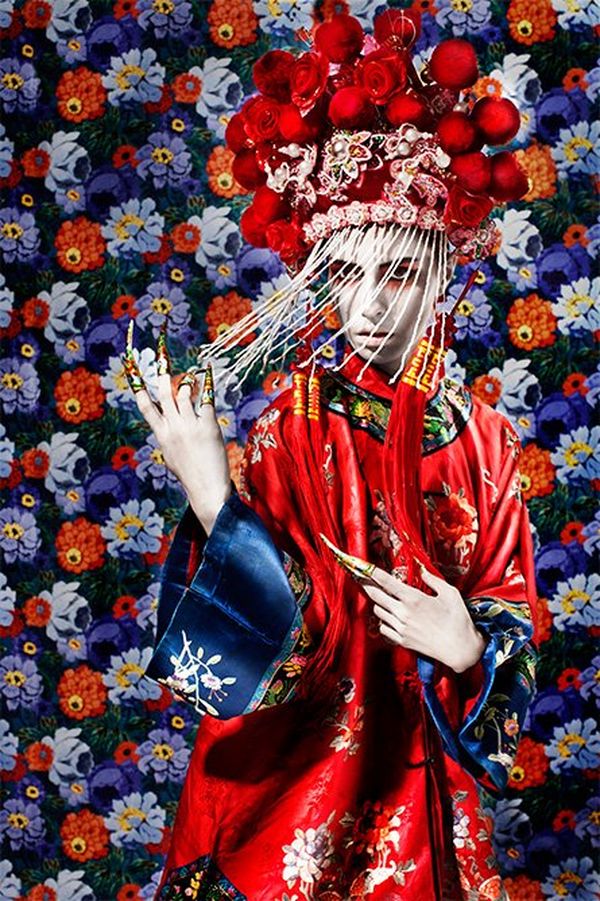 Wearing an ornate red and blue satin kimono and red headdress, a woman stands against a background of richly-coloured flowers.