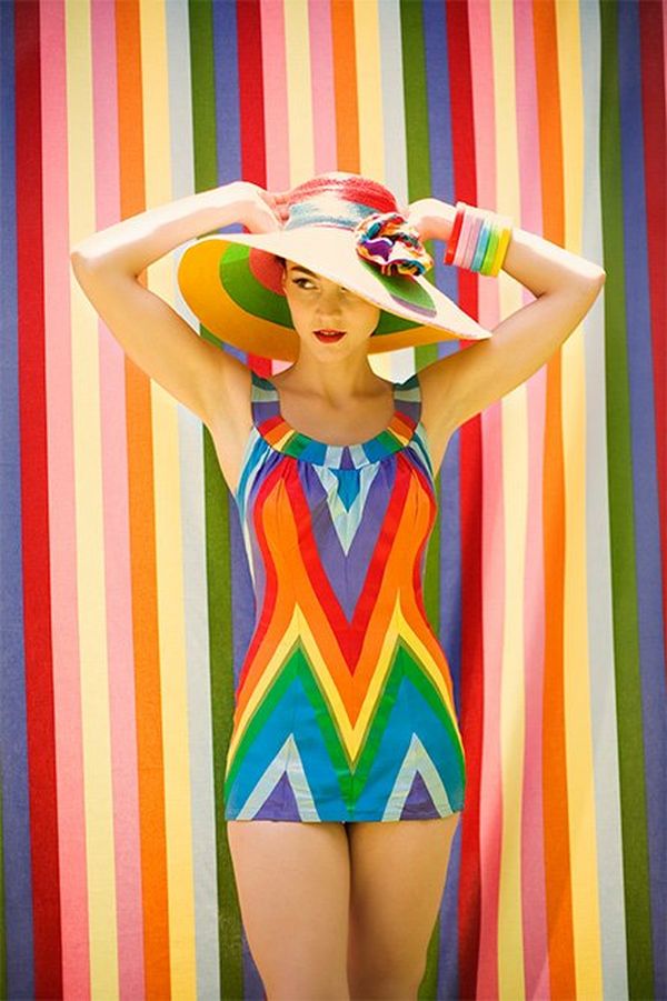 A woman in a rainbow-coloured, stripy, retro-style swimsuit stands against a matching wall, holding a matching sun hat to her head.