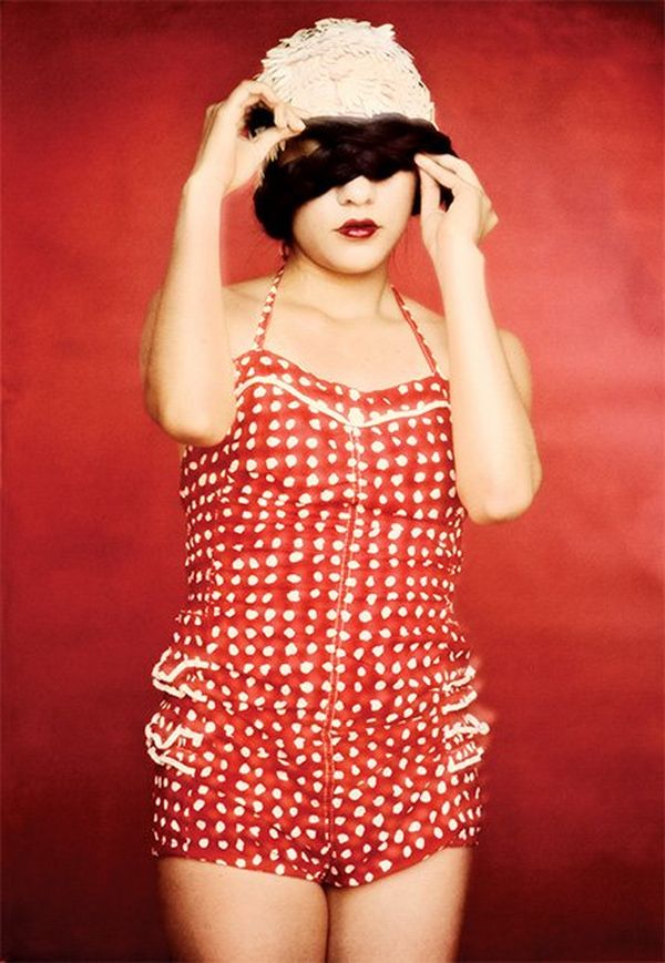 A model wears a vintage-style red-and-white swimsuit and a white swimming cap. She holds a black blindfold on her face.
