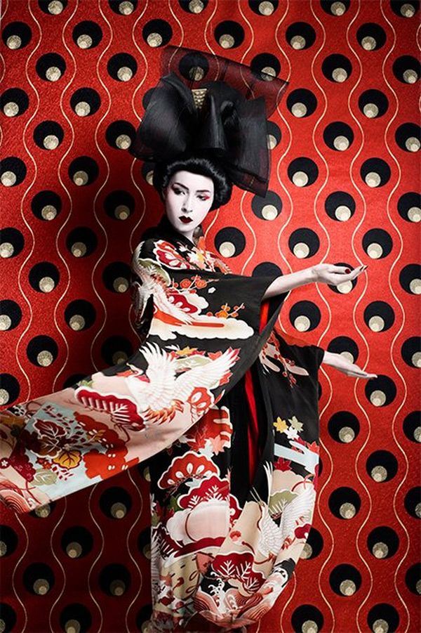 A model is dressed as a Japanese woman in an ornate red-and-white kimono and black headdress, standing before a richly patterned wall, the predominant colours of which are red, gold and black.