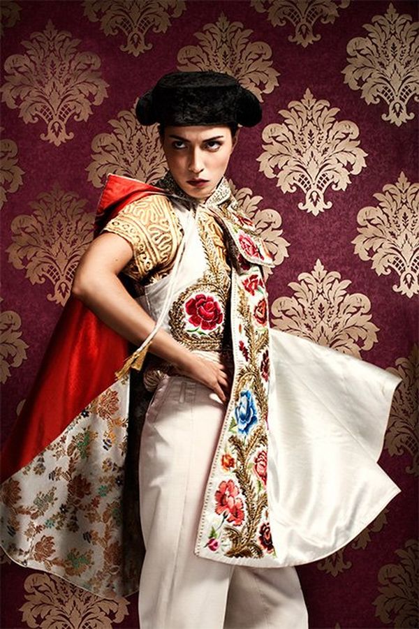 A model is dressed like a matador with ornate satin clothing and hat, standing before a red and gold brocade background.