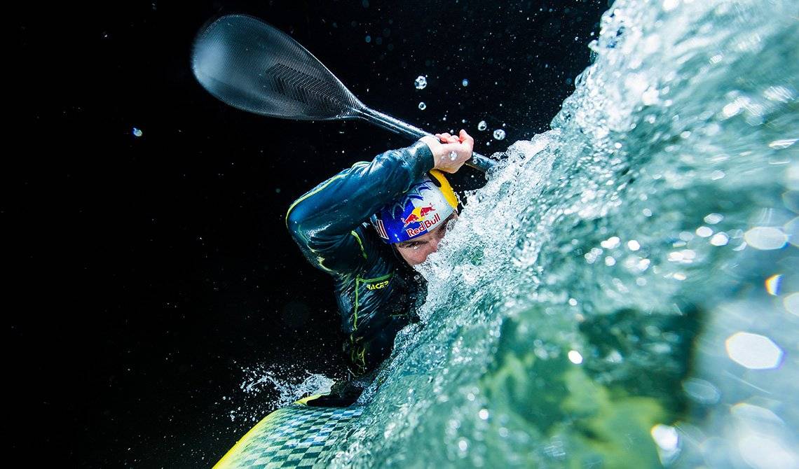 Kayaker Peter Kauzer leans to one side as he paddles through white water, spray highlighted against the night sky.