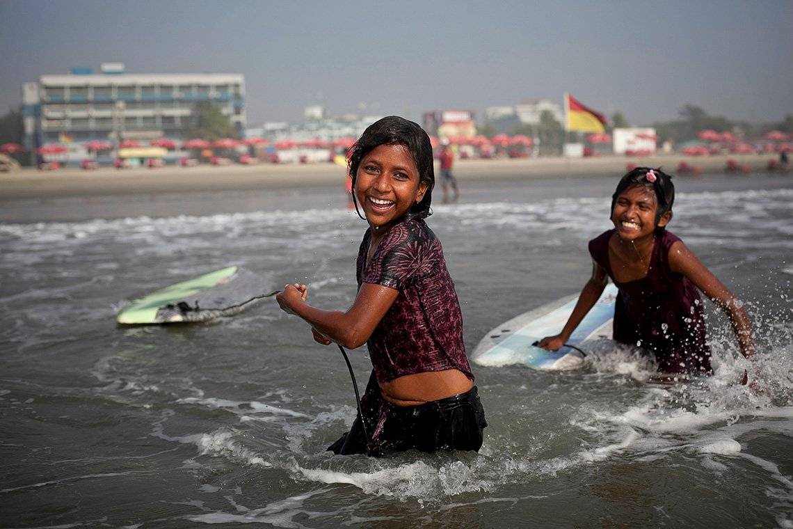 Two 10-year-old Bangladeshi girls smile as they drag their surfboards into the sea, with the beachfront and hotels of Coxs Bazar in the background.