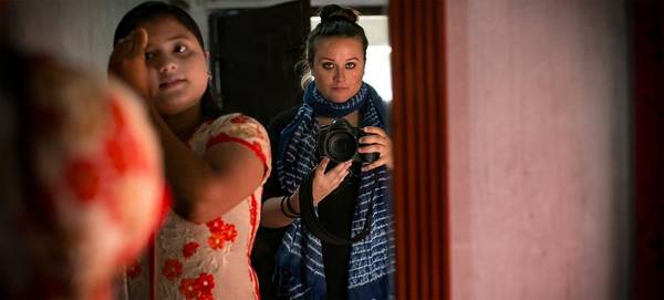 American photographer Allison Joyce takes a photograph of herself in a mirror, holding a Canon DSLR, next to a young Asian girl who is brushing her hair.