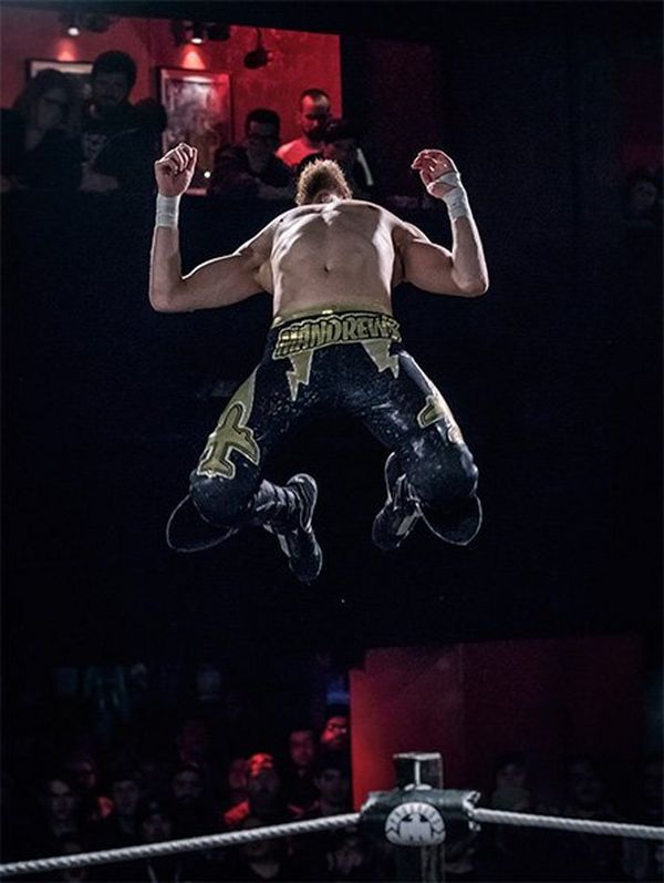 A wrestler jumps high in the air, head back. The crowd look on behind him.