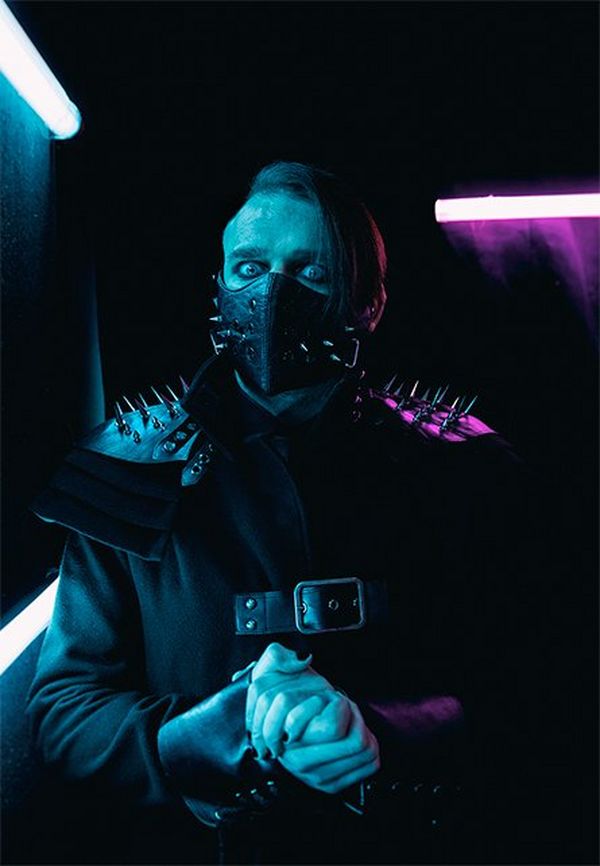Wrestler Jimmy Havoc wears a studded black leather mask over his mouth, and a black outfit with spiked shoulder pads, as he looks at the camera.
