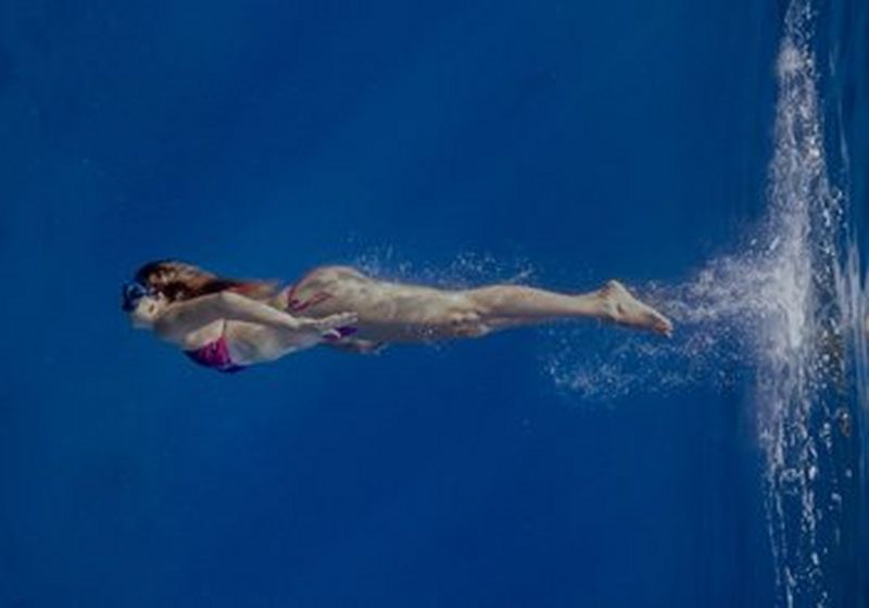 A woman in a pink bikini and goggles swims through royal blue water. To her right are the bubbles that signify impact into the water and her reflection further beyond them.