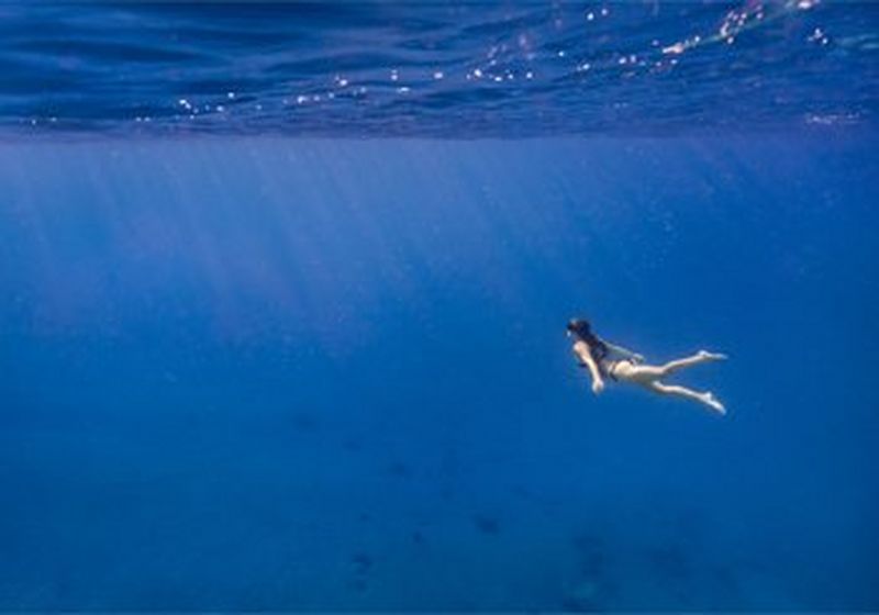 An image taken underwater. The sea is bright blue and a woman in a bikini and snorkel swims upwards towards the light and bubbles on the surface. © Dafna Tal