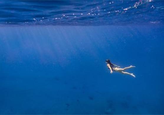 An image taken underwater. The sea is bright blue and a woman in a bikini and snorkel swims upwards towards the light and bubbles on the surface. © Dafna Tal