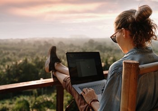 A young woman in sunglasses sits outside on a wooden chair with her laptop on her lap. Her feet are resting on a wooden beam that appears to be enclosing a balcony or raised veranda. The view takes in a vast expanse of greenery and there are hills in the distance.  