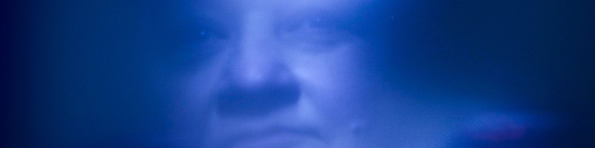 A clouded and slightly blurred image, all in blue. Through it, the features of a woman’s face can be seen. © Daphne Wageman