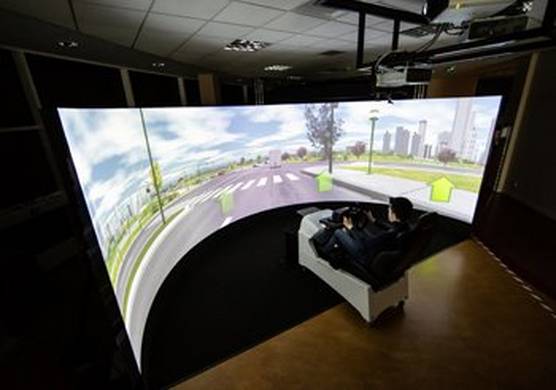 A person sits in a simulator unit in a darkened room, holding a steering wheel. In front of them is a huge, curved screen upon which a driving scene is projected.