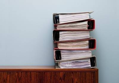 A stack of ring binders full of papers piled on top of one another on a mahogany filing cabinet.