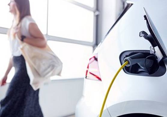 A shot of the rear of a white electric car being charged using a charger with a yellow cable. In the background, a blurred long-haired person in a blue skirt and white top walks away, carrying a cream-coloured bag over their shoulder.