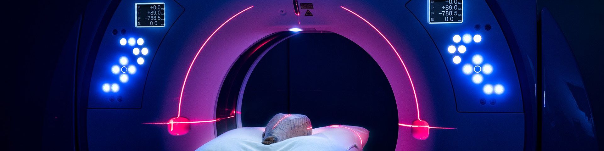 An object lies on a bed, inside a round CT scanner. It is illuminated with blue and purple lights.