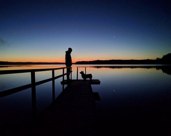 A person stands with their dog on decking over water. The sun is setting in the background in a beautiful orange and the sky is a deep blue.