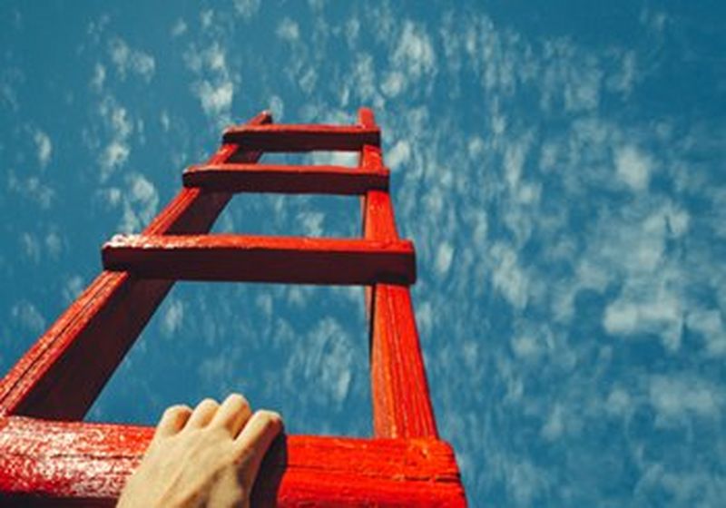 A hand reaching onto a red ladder, which points at a bright blue sky