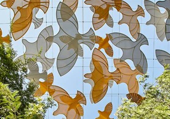 Cut out orange and grey shapes of birds, against a blue sky, with trees on either side. This is the view from beneath of a section of the Terra Sustainability Pavilion at Expo Dubai 2020.