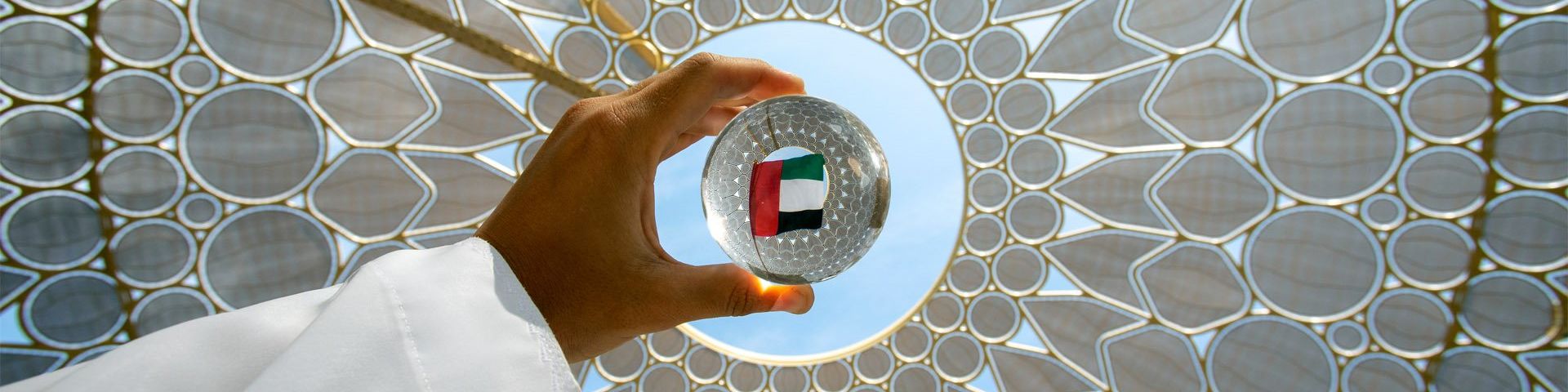 A white sleeved arm holds a glass ball against the sky light through a circular hole in a white ceiling of geometric design. In the centre of the glass ball, the flag of the UAE can be seen.