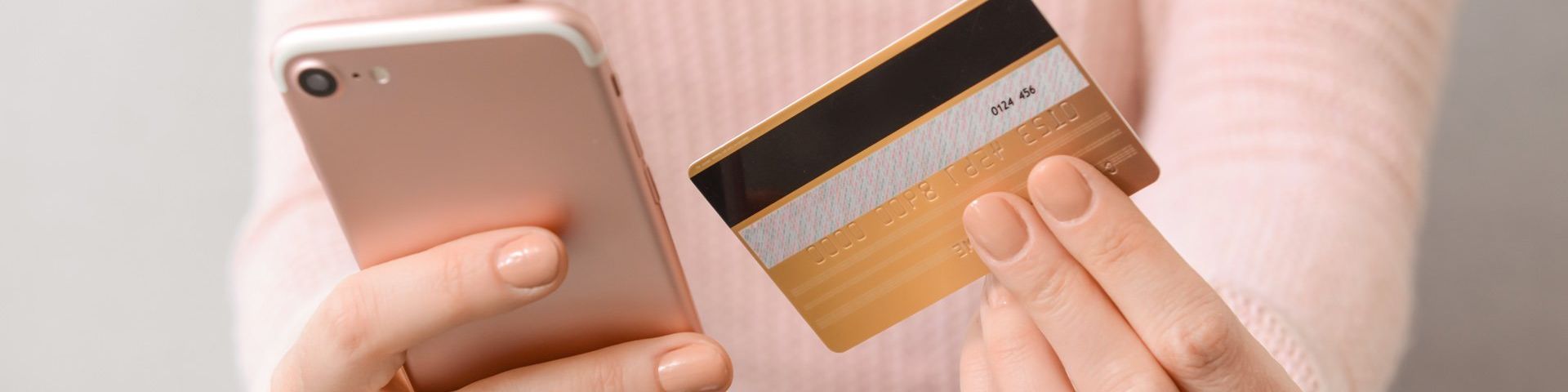 An unseen person in a pale pink sweater holds a pink smartphone in one hand and a credit card in the other.