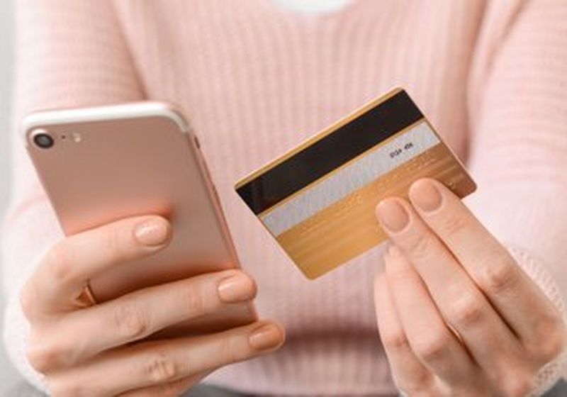 An unseen person in a pale pink sweater holds a pink smartphone in one hand and a credit card in the other.