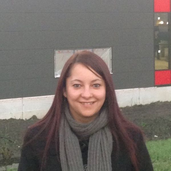 A smiling, brown haired woman stands in front of a building, wearing a black coat and grey scarf.