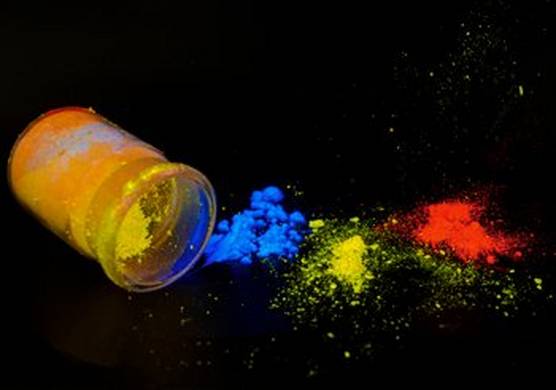 On the left is a small glass bottle on its side. It contains bright yellow powder. Beside it to the right are small piles of bright blue, yellow and red powder. This is all against a black background.