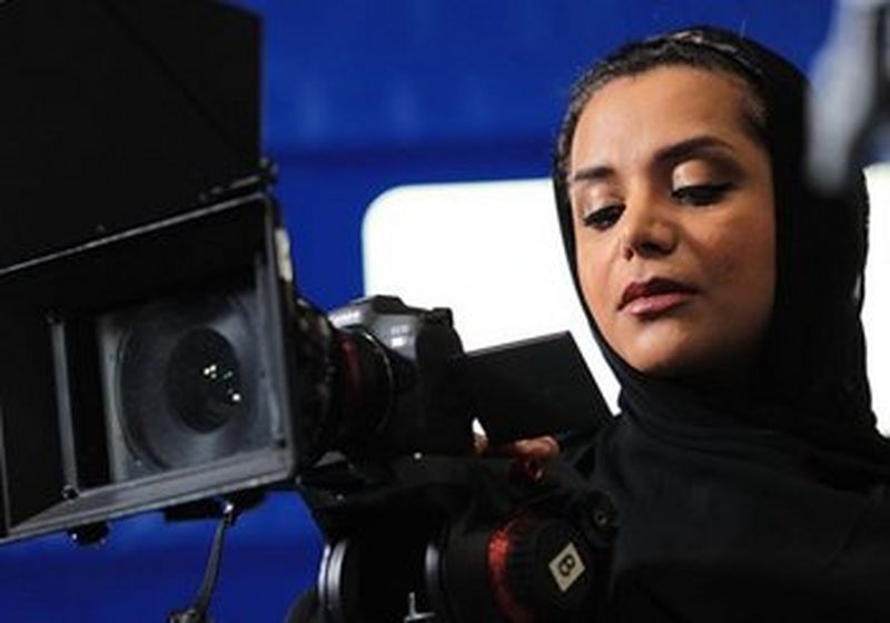 Filmmaker & Associate Producer, Nayla Al Khaja. She holds a film camera and stands against a blue background.