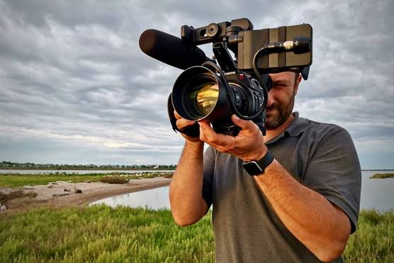 Filmmaker Roberto Palozzi filming with a Canon video camera with his back to the water's edge. 