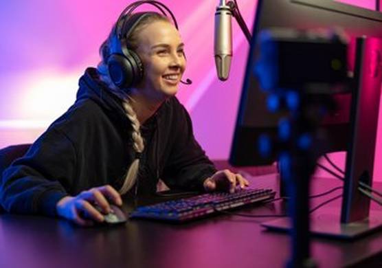 A smiling young woman with her hair in a long blonde braid sits at a desktop computer with keyboard. She wears over-ear headphones and has a big silver microphone positioned in front of her.