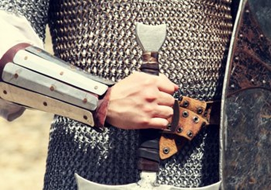 A knight’s chain-mailed torso and arm, with the handle of a sword and partial shield showing.