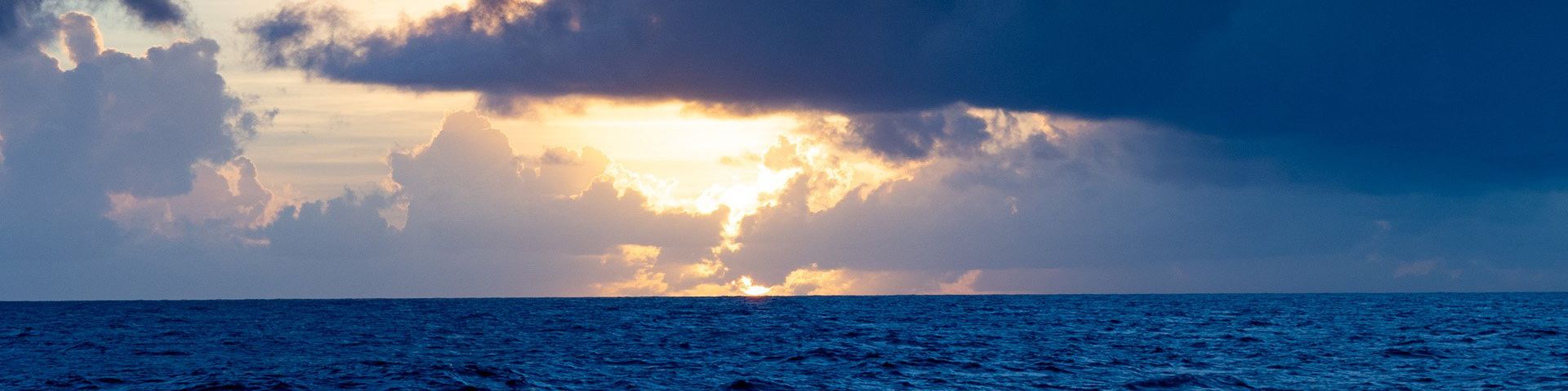 The sun shines from behind clouds above a dark blue ocean (© Richard Garriott de Cayeux, using Canon EOS R / Canon RF24-105mm F4 IS USM)