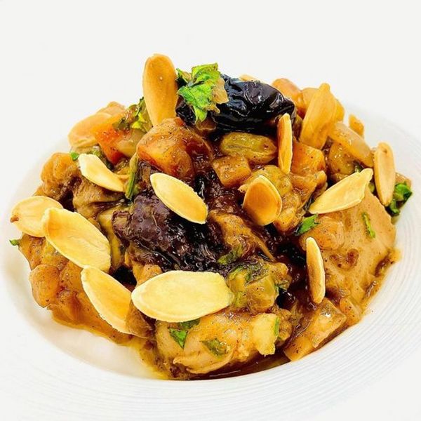 The golden-brown tagine sits in a white bowl on a white background. It is garnished with almonds and a green leaf.