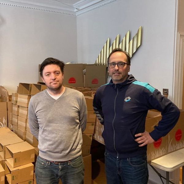 Two men in casual clothes stand in front of stacks of brown delivery boxes.