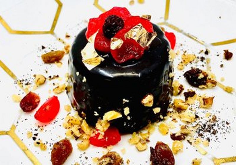 A dark brown chocolate dessert that shines under the light is topped with gold flakes, berries and nuts. It sits on a white plate with a geometric gold honeycomb design.