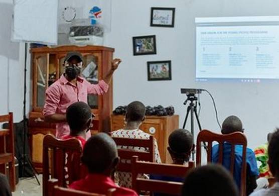 A man stands points at a projector screen, as he teaches a class of young people.