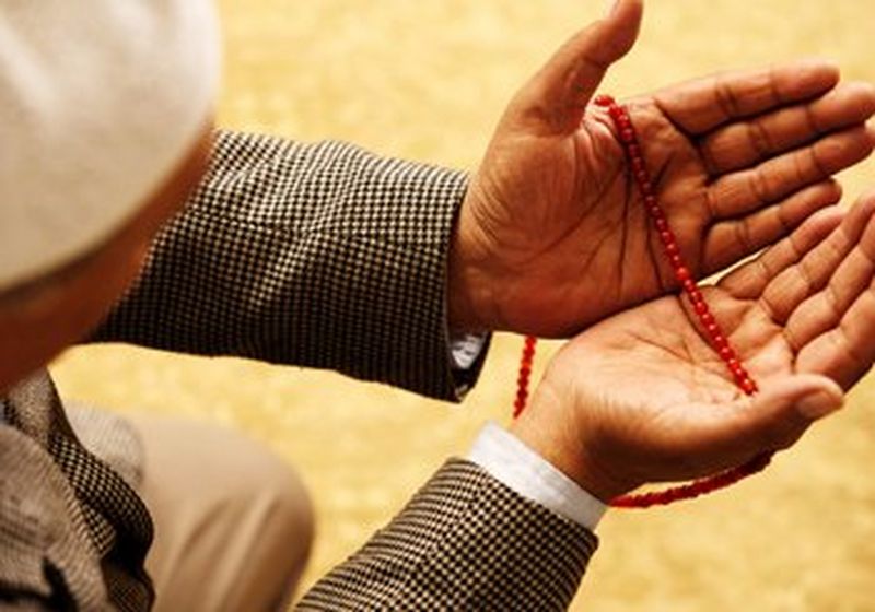 Viewed from above, a man holds his hands out in front of him in prayer. In them are red prayer beads. From his arms we can see that he is wearing a checked suit jacket and a white shirt.