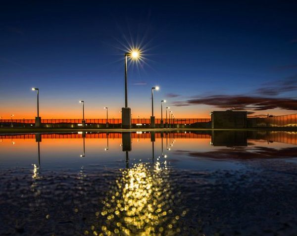 A dark blue sky is reflected over water and the light from lampposts is scattered over the water. Where the water meets the sky in the distance the sun is setting in orange hues.