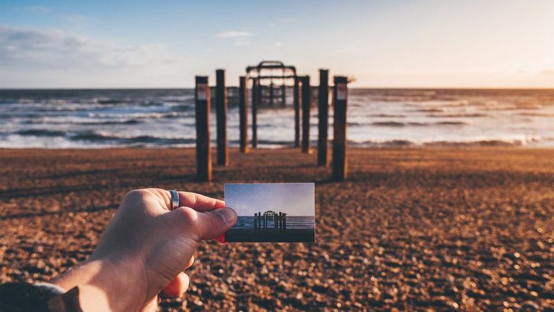 Joel Thorpe holding a print on Brighton beach, in front of the remains of the old pier. 