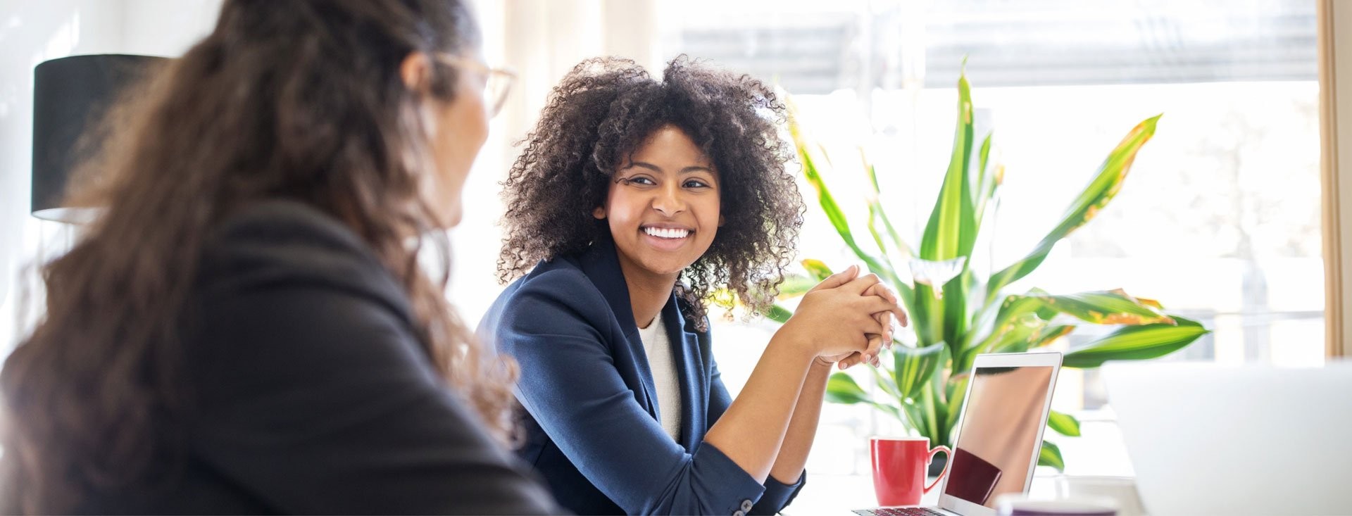 Woman smiles at a colleague while both sat at a white desk with a bright green plant