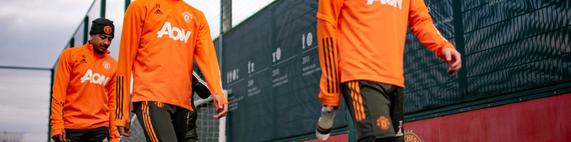 Three footballers walk in line, in front of a black and red barrier. Only one of their faces is shown. They are wearing bright orange shirts with ‘AON’ printed on the front and black shorts with orange stripes down the outer leg. Photo courtesy of Manchester United FC.