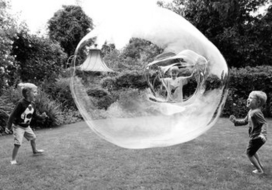Three children in a garden, one is making a huge soap bubble from a hoop. The others are laughing and screaming with the fun of it all.