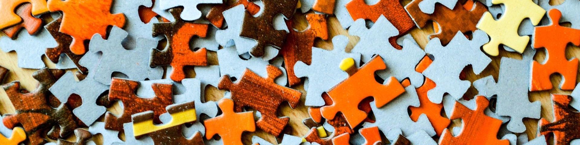 The pieces of a jigsaw puzzle, orange and brown pieces, mixed up with others that are face down, showing only the grey cardboard on their reverse.