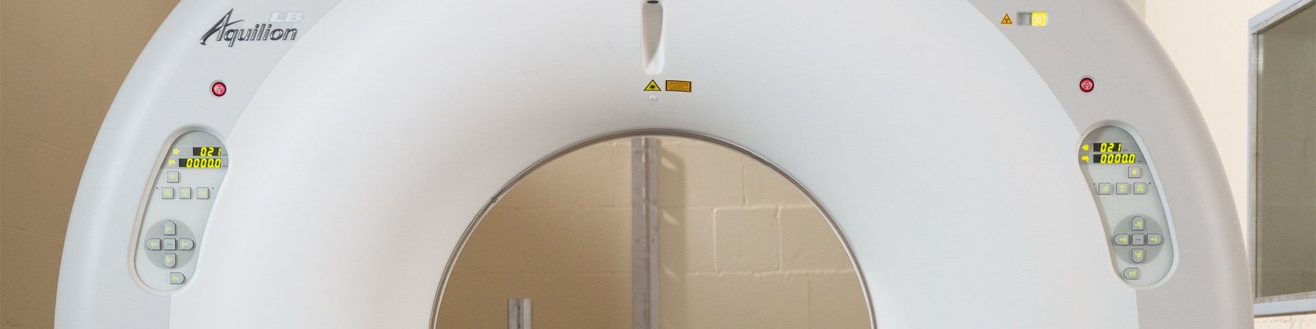 A white, half-doughnut shaped machine, which is a partial view of a Canon Medical Aquilion LB CT Scanner. It has operating buttons on either side of the central gantry.